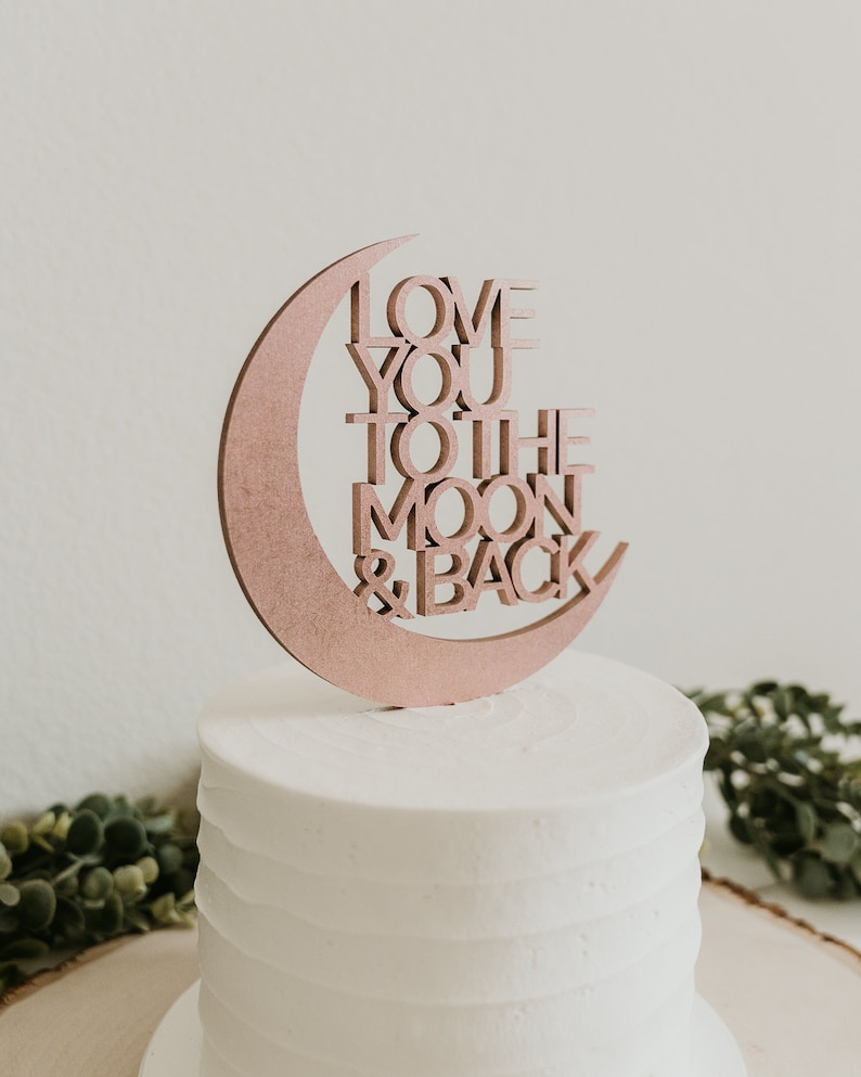 May include: A white cake with a wooden cake topper in the shape of a crescent moon with the words "Love You To The Moon & Back" written on it.