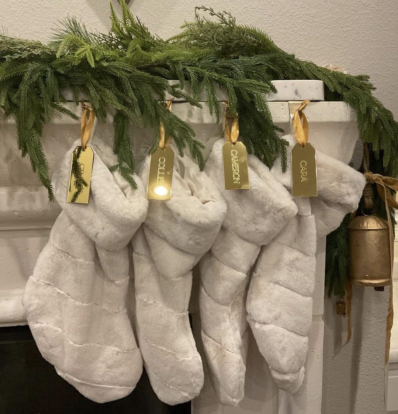 Op de afbeelding: Vier witte kerstsokken van imitatiebont hangen op een rij, elk voorzien van een goudkleurig metalen label bevestigd met een gouden lint. De labels zijn gegraveerd met de namen Colleen, Cameron en Cara. Boven de sokken hangt een groene dennenkrans.