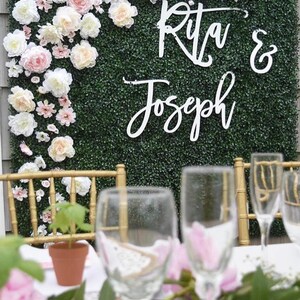 May include: A green artificial hedge backdrop decorated with white and pink flowers. The backdrop has a white sign with the text "Rita & Joseph" in cursive.
