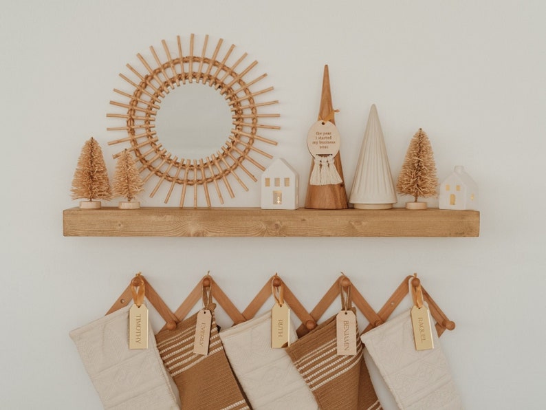 Op de afbeelding: Een houten wandplank toont een zonnespiegel, twee witte keramische huisjes, twee flessenborstelbomen en twee decoratieve kegels. Daaronder hangt een accordeonrek met vijf kousen voorzien van gepersonaliseerde gouden naamplaatjes.