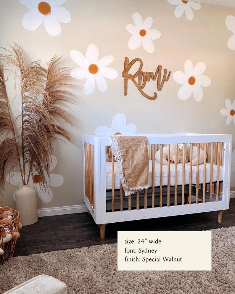 Puede incluir: Una cuna blanca con un marco de madera y una alfombra marr&oacute;n debajo. La pared detr&aacute;s de la cuna est&aacute; pintada de color beige claro y presenta flores de margarita blancas con centros naranjas. El nombre "Romu" est&aacute; escrito en cursiva en la pared con una fuente de madera. El texto debajo de la cuna dice "tama&ntilde;o: 24" de ancho, fuente: Sydney, acabado: Nogal Especial".