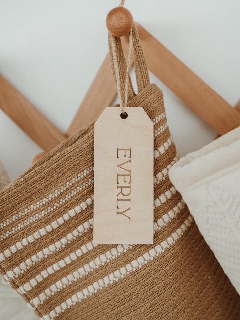 Op de afbeelding: Een rechthoekig, lichtgekleurd houten cadeaulabel met de naam EVERLY in hoofdletters gegraveerd. Het label is met een dun touwtje van jute bevestigd aan een gestructureerde, beige en witte geweven kous die aan een houten haak hangt.