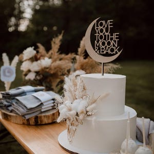Puede incluir: Un pastel de boda blanco de dos niveles decorado con flores secas y un adorno de pastel de madera que dice "Love you to the moon & back".
