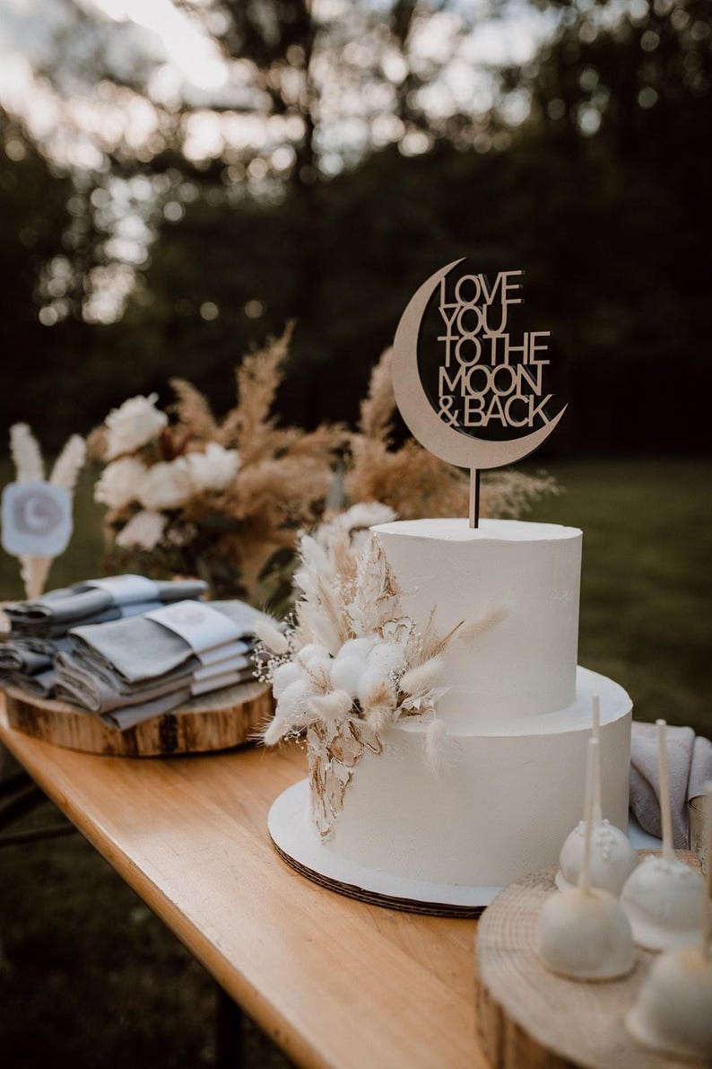 Puede incluir: Un pastel de boda blanco de dos niveles decorado con flores secas y un adorno de pastel de madera que dice "Love you to the moon & back".