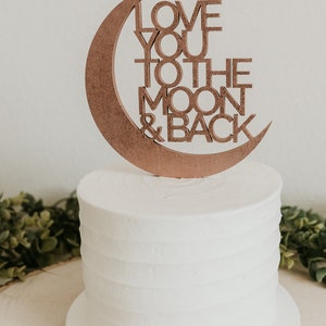 May include: A white cake with a wooden moon and back topper that says "Love you to the moon & back".