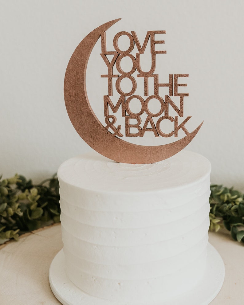 May include: A white cake with a wooden moon and back topper that says "Love you to the moon & back".
