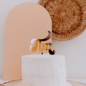 May include: A white cake with a gold "Tim" cake topper on top. The cake is on a wooden cake stand with a brown fan on the side.