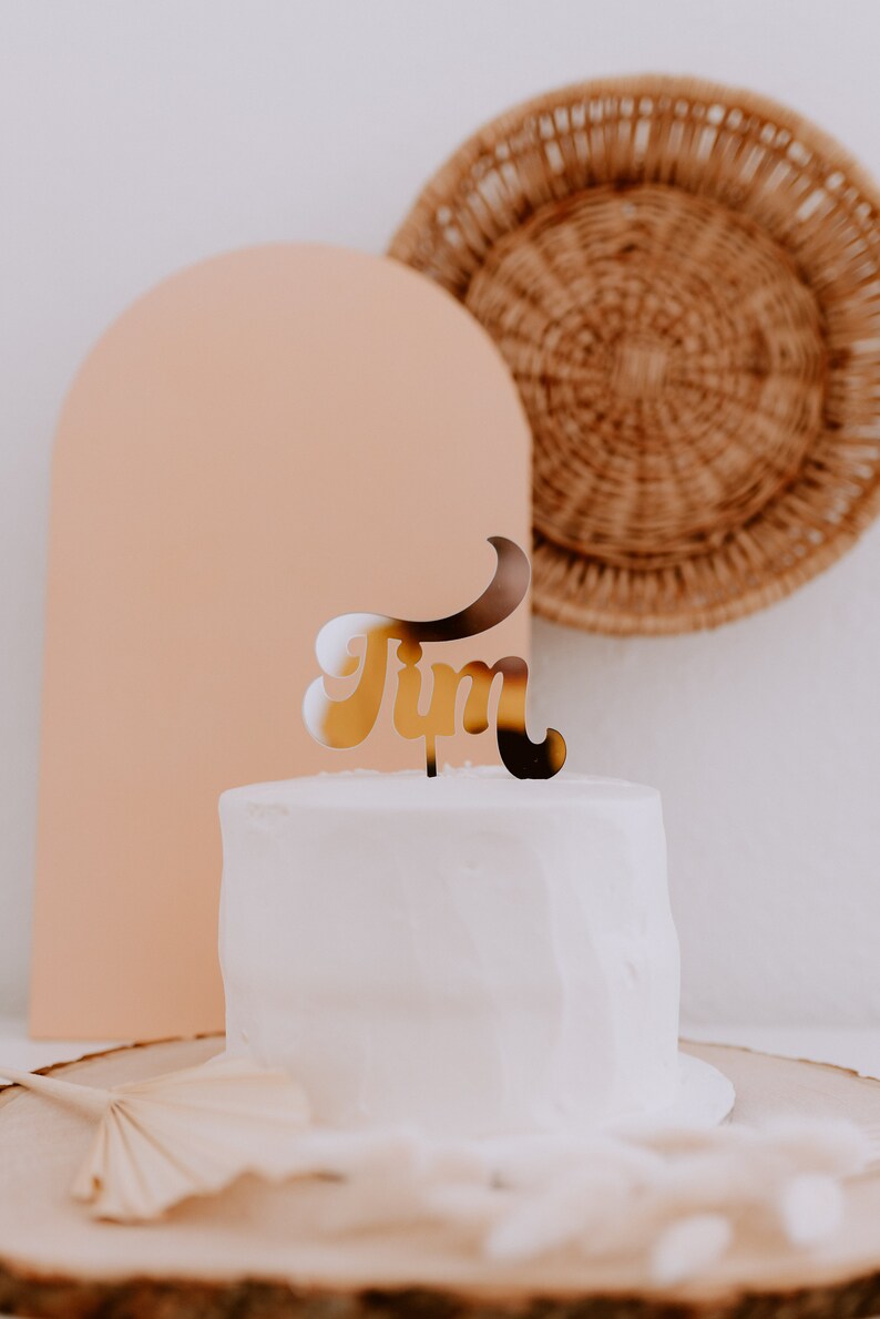 May include: A white cake with a gold "Tim" cake topper on top. The cake is on a wooden cake stand with a brown fan on the side.
