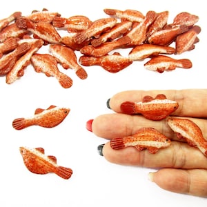 Peut inclure: Collection de petites figurines en forme de poisson, orange et blanc. Les figurines sont détaillées avec des nageoires et des écailles, et certaines sont tenues dans une main. Les poissons sont dispersés sur une surface blanche.