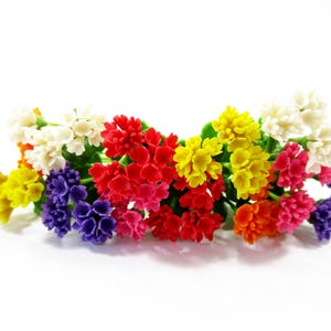 May include: A vibrant arrangement of colorful artificial flowers. The bouquet features clusters of small blossoms in shades of white, yellow, red, pink, purple, and orange, set against green foliage. The flowers are arranged on a white background.