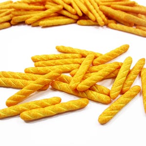 May include: A close-up shot of golden-yellow, breadstick-shaped snacks with a textured surface. The snacks are scattered on a white surface, with some arranged in a pile in the background. The snacks have a slightly orange hue.