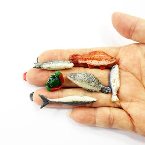 May include: A collection of miniature fish figurines in various colors, including red, white, gray, and blue. The figurines are arranged in a person's hand against a white background. The fish vary in shape and size.
