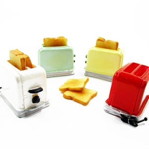 Op de afbeelding: Vier miniatuurtoasters in wit, groen, geel en rood, elk met een sneetje toast. Twee extra sneetjes toast liggen voor de toasters. De toasters staan op een witte ondergrond.
