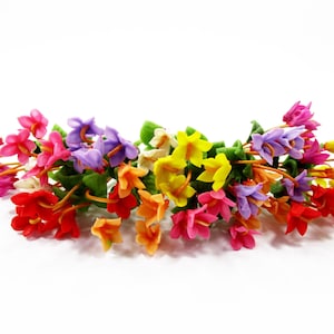 Puede incluir: Un arreglo floral decorativo que presenta un grupo de pequeñas flores multicolores en tonos rosa, morado, amarillo, naranja y rojo. Cada flor tiene tallos delgados y pequeñas hojas verdes, creando un ramo denso y vibrante de flores individuales.