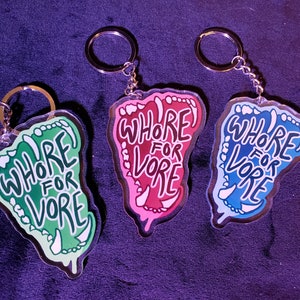 May include: Three acrylic keychains shaped like a mouth with teeth. Each keychain has the text "WHORE FOR VORE" written inside the mouth. The keychains are green, pink, and blue.