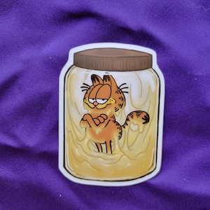 May include: A die-cut sticker featuring a cartoon orange tabby cat with black stripes and a grumpy expression, submerged inside a clear glass jar filled with a thick, pale yellow substance, topped with a brown screw-on lid and a white border.