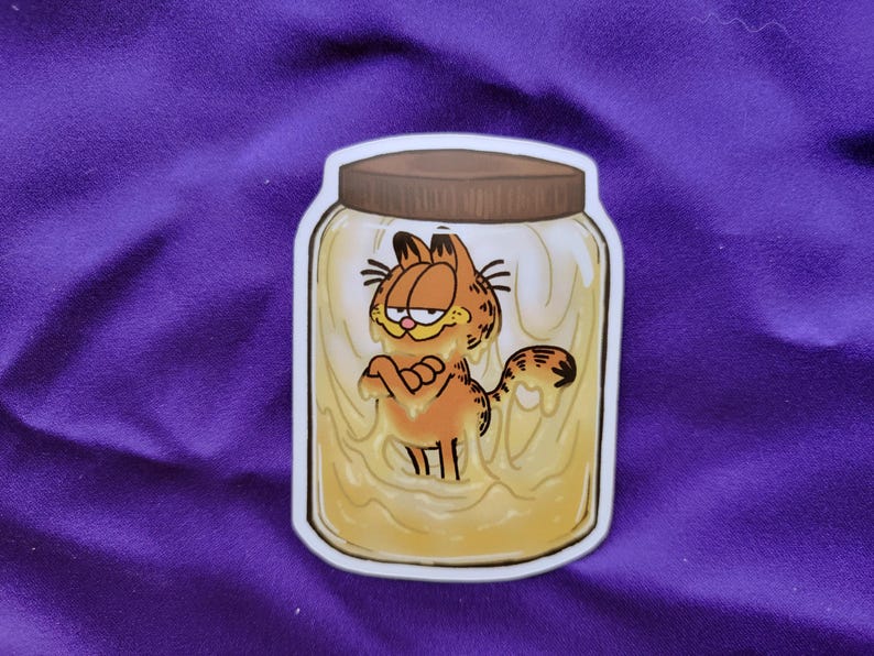 May include: A die-cut sticker featuring a cartoon orange tabby cat with black stripes and a grumpy expression, submerged inside a clear glass jar filled with a thick, pale yellow substance, topped with a brown screw-on lid and a white border.