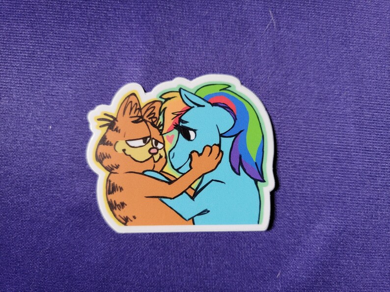 May include: A die-cut sticker featuring a cartoon orange tabby cat with black stripes embracing a light blue pony with a multicolored rainbow mane. The cat has a smug expression, and the pony has long eyelashes and a small pink heart between them.