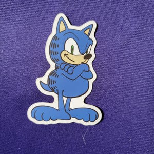 May include: A die-cut sticker featuring a blue cartoon character with a round body, large feet, and a tail, wearing a blue hood with pointed ears that resembles the head of a hedgehog. The character has a tan face, green eyes, and crossed arms.