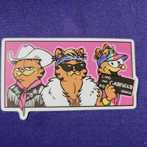 May include: A rectangular sticker on a purple background featuring three orange tabby cats in a pink frame. The left cat wears a white cowboy hat and fringed shirt. The middle cat wears sunglasses, a bandana, and a white fur coat. The right cat wears a colorful visor and holds a sign reading LAPD AND GARFIELD 919 VENICE.