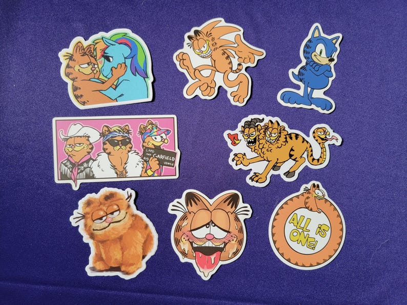 May include: A collection of eight vinyl stickers featuring various illustrations of the orange tabby cat Garfield. Designs include Garfield with a blue pony, as Sonic the Hedgehog, in a cowboy hat, as a multi-headed creature, and text reading All is one.