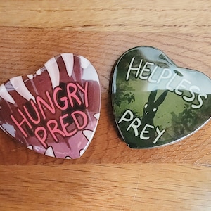 May include: Two heart-shaped pins with contrasting designs. One pin is red with white stripes and says "HUNGRY PRED" in pink and white lettering. The other pin is green with a black rabbit silhouette and says "HELPLESS PREY" in white lettering.