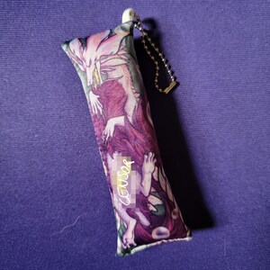 May include: A rectangular, plush keychain featuring a fantasy illustration in shades of purple, pink, and white. The artwork depicts a mythical creature and a figure. The keychain has a silver chain and a white plastic attachment.