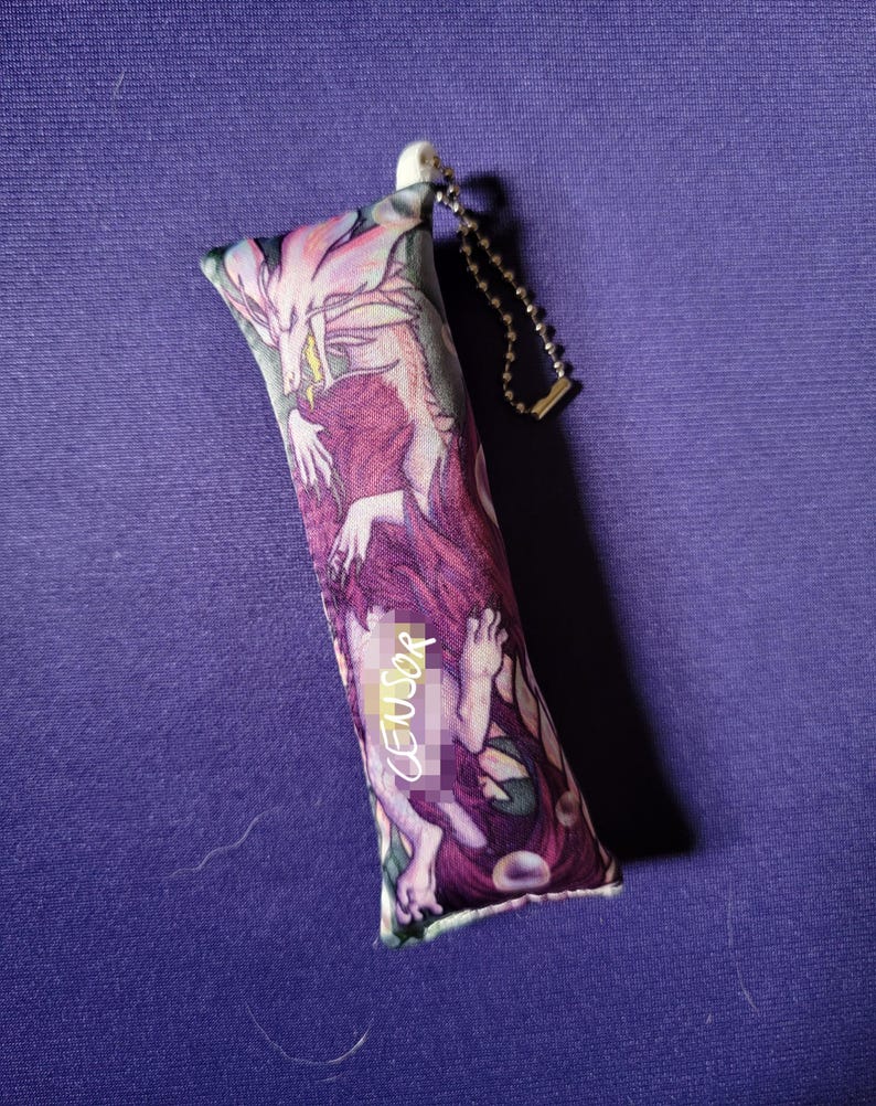 May include: A rectangular, plush keychain featuring a fantasy illustration in shades of purple, pink, and white. The artwork depicts a mythical creature and a figure. The keychain has a silver chain and a white plastic attachment.