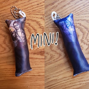 Puede incluir: Dos pequeñas almohadas de felpa con llaveros. Una almohada es azul oscuro con un diseño blanco y azul y la otra es negra con un diseño marrón y blanco. El texto "MIN!" está escrito en blanco sobre fondo negro.