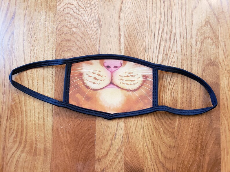 May include: A black fabric face mask with a printed image of a cat's nose and whiskers. The cat's fur is orange and white.