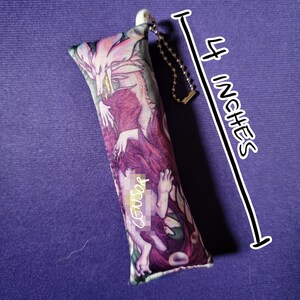May include: A rectangular, fabric item with a fantasy art print, approximately 4 inches long. The artwork features a dragon and figures in shades of purple, pink, and white. A small chain and white plastic piece are attached to the top. The word "CENSOR" is visible on the item.