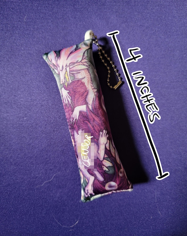 May include: A rectangular, fabric item with a fantasy art print, approximately 4 inches long. The artwork features a dragon and figures in shades of purple, pink, and white. A small chain and white plastic piece are attached to the top. The word "CENSOR" is visible on the item.