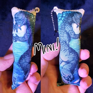 May include: A blue and green plush keychain featuring a printed image of a monster. The keychain is shaped like a pillow and has a small loop for attaching to a bag or keys. The text "Mini!" is written in black on a white background.