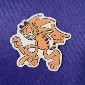 May include: A die-cut sticker featuring a mashup character design of an orange tabby cat with black stripes and a spiky hairstyle, posed in a dynamic running stance with a wide, toothy grin, set against a solid purple fabric background.