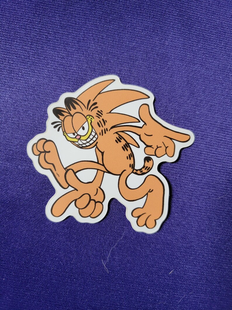 May include: A die-cut sticker featuring a mashup character design of an orange tabby cat with black stripes and a spiky hairstyle, posed in a dynamic running stance with a wide, toothy grin, set against a solid purple fabric background.
