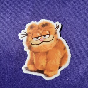 May include: A die-cut sticker featuring a fluffy orange tabby cat with dark brown stripes, a small pink nose, and a smug expression with half-closed eyes. The illustration has a soft, textured appearance with visible whiskers and a white border.