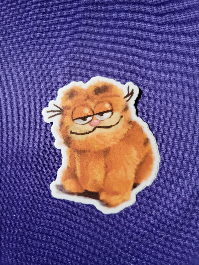 May include: A die-cut sticker featuring a fluffy orange tabby cat with dark brown stripes, a small pink nose, and a smug expression with half-closed eyes. The illustration has a soft, textured appearance with visible whiskers and a white border.