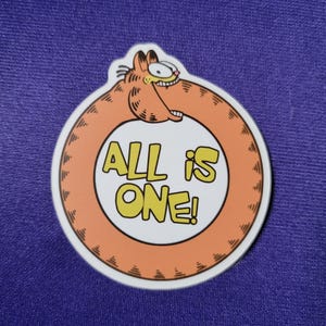 May include: A circular sticker featuring a cartoon orange cat biting its own tail to form a ring. The center is white with the yellow text ALL IS ONE! in a bold, blocky font. The cat has black stripes, a wide grin, and visible teeth.