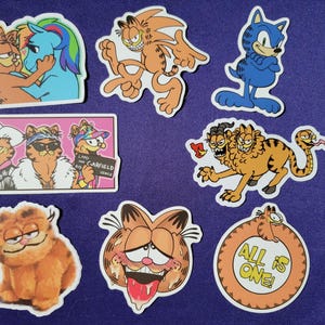 May include: A collection of seven die-cut vinyl stickers featuring various cartoon cat illustrations, including a multi-headed creature, a cat-pony hybrid, and a circular design with the text ALL IS ONE, displayed next to a white measuring tape showing 8 inches.