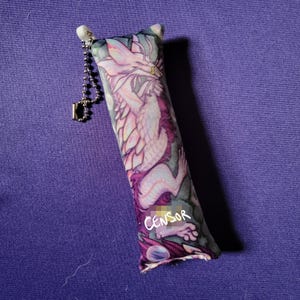 May include: A rectangular cat toy with a dragon illustration in shades of pink, purple, and white. The toy has a silver chain and a small tag. The word "CENSOR" is written on the toy.