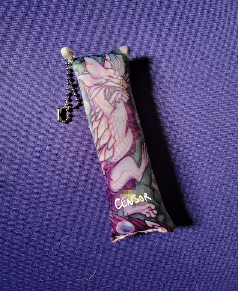 May include: A rectangular cat toy with a dragon illustration in shades of pink, purple, and white. The toy has a silver chain and a small tag. The word "CENSOR" is written on the toy.