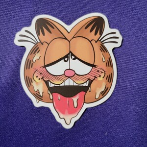 May include: A die-cut sticker featuring a stylized orange cartoon cat face with black stripes, large white eyes with small hearts, a pink nose, flushed cheeks, and an open mouth with a long pink tongue hanging out, surrounded by a white border.