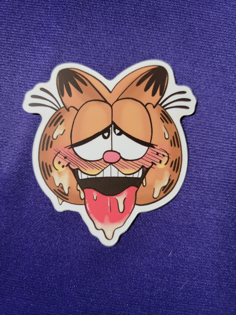 May include: A die-cut sticker featuring a stylized orange cartoon cat face with black stripes, large white eyes with small hearts, a pink nose, flushed cheeks, and an open mouth with a long pink tongue hanging out, surrounded by a white border.
