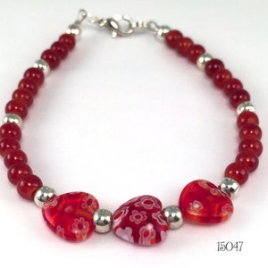 May include: Red beaded bracelet with three red heart-shaped charms with white floral designs. The bracelet has a silver clasp and silver beads.