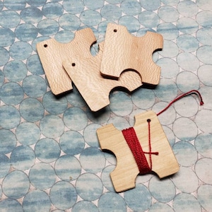 May include: Four wooden thread bobbins with a red thread wound around one of them. The bobbins have a unique shape with a hole in the center and a notch on each side.