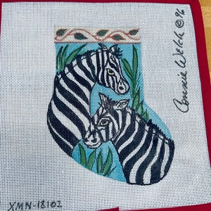 May include: A white canvas needlepoint stocking with a red border. The stocking features a blue background with two zebras, one larger than the other, and green foliage. The text "Connie Wilson" is written in black on the right side of the stocking.