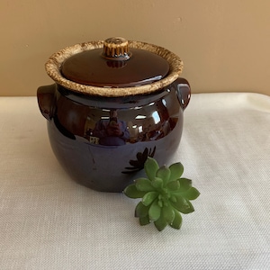 May include: A brown glazed ceramic crock with a lid. The crock has a brown and tan glaze with a mottled pattern. The lid has a knob on top.
