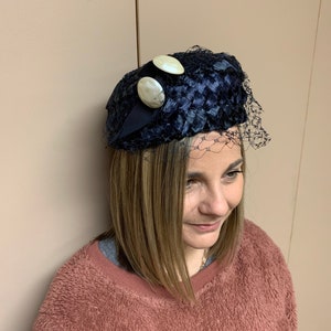 May include: A navy blue velvet hat with a netting veil and two white buttons. The hat is worn by a person with brown hair.