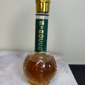 May include: A small, round glass bottle with a gold cap and a green label that says "SIROCCO". The bottle is filled with a brown liquid.
