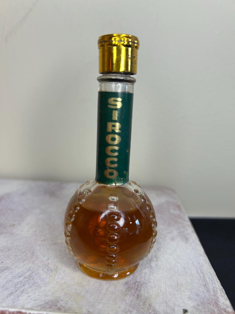 May include: A small, round glass bottle with a gold cap and a green label that says "SIROCCO". The bottle is filled with a brown liquid.